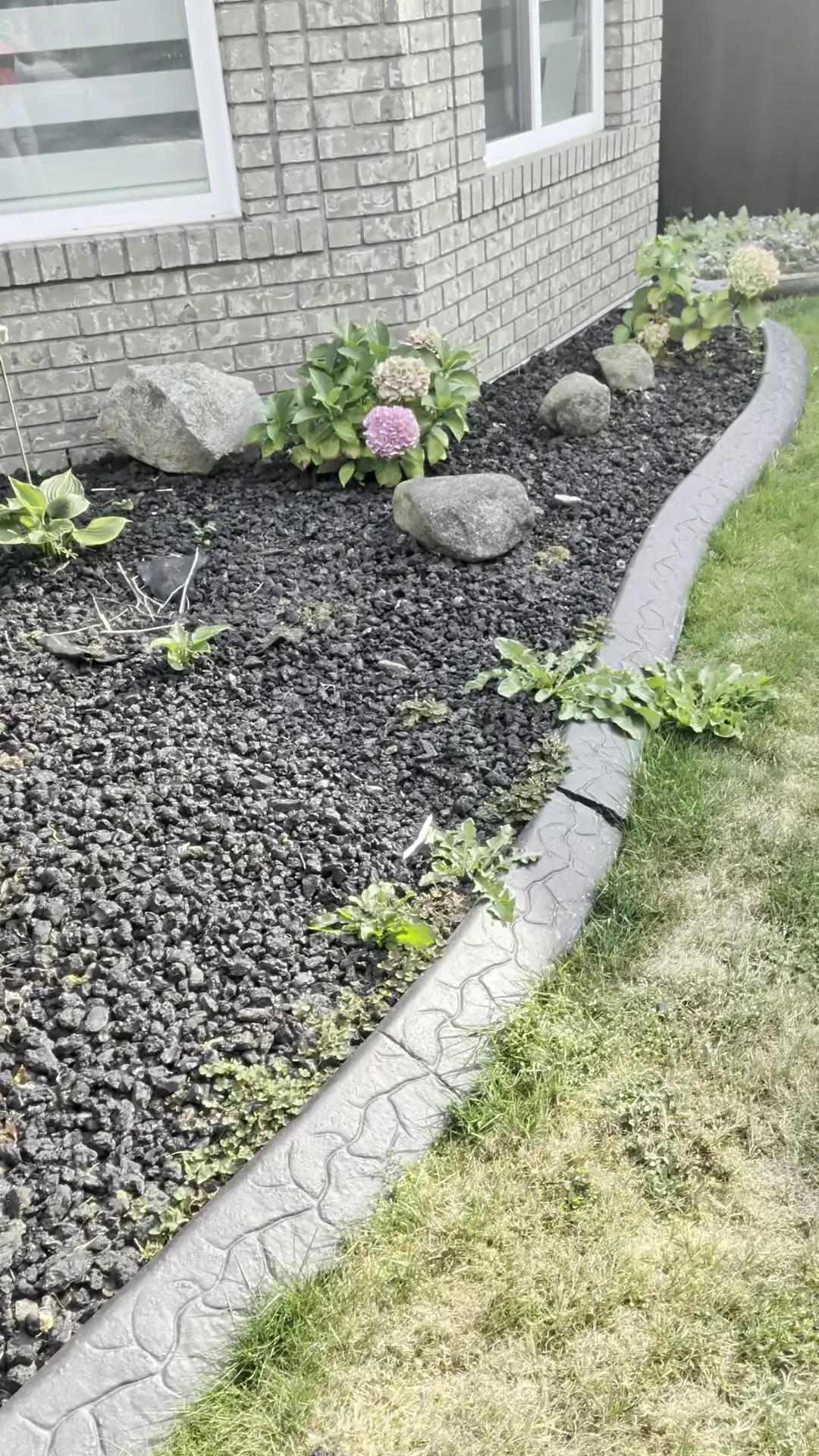 Rock garden bed against a house wall, faded and dotted with weeds, before refresh