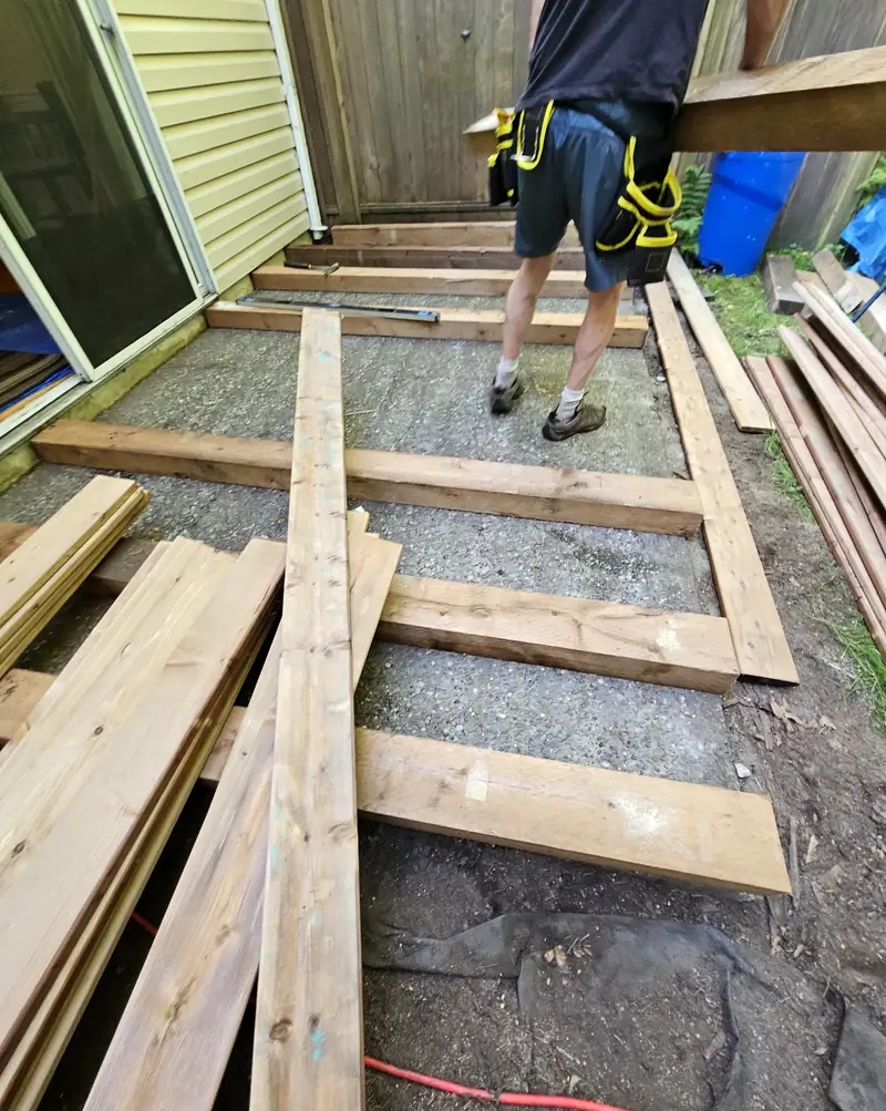An Ironwood Grounds crew member laying out pressure-treated deck framing on a gravel base, with cedar deck boards stacked alongside