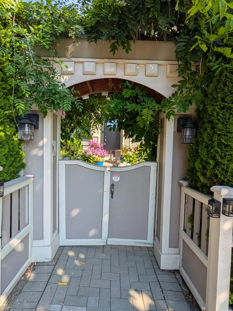 Street side of an arched arbor entry before, with an old painted grey double gate on a paver path